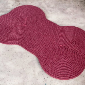 May include: A pink, woven, bone-shaped rug with a brown leather tag.