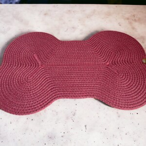 May include: A red, woven, bone-shaped pet mat. The mat is made of thick, braided rope and has a slightly textured surface.