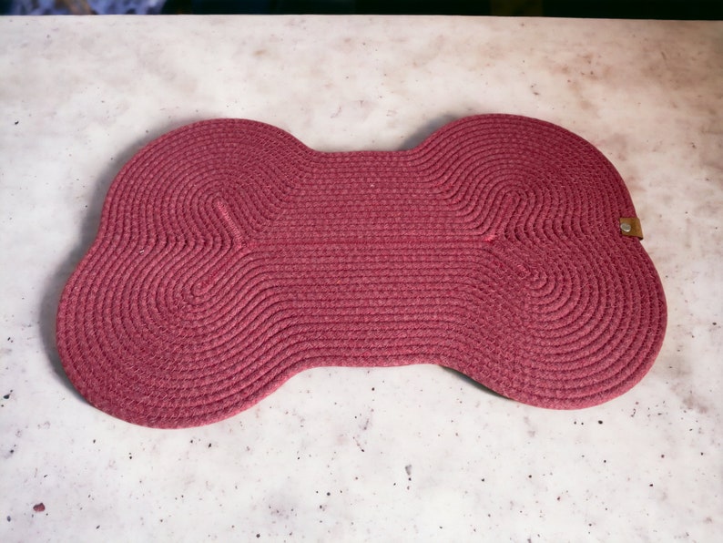 May include: A red, woven, bone-shaped pet mat. The mat is made of thick, braided rope and has a slightly textured surface.