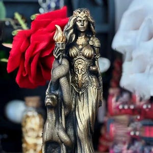 Può includere: Statua color bronzo di una donna che tiene una torcia e un serpente, con un cane ai suoi piedi. La statua presenta dettagli intricati, tra cui una corona e abiti ornati. La base della statua presenta testo greco.