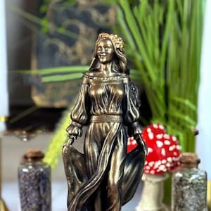 May include: Bronze-colored statue of a woman in a flowing gown, holding the fabric. She wears a floral crown and stands on a base with text. The statue is on a wooden surface, with jars and decorative elements in the background.