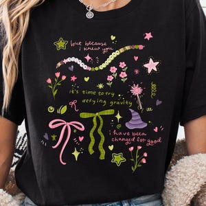 May include: Black t-shirt with a graphic print of a witch's hat, stars, bows, and the text "but because I knew you it's time to try defying gravity I have been changed for good".