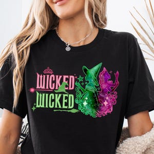 May include: Black t-shirt with the words "Wicked Wicked" in pink and green, and a graphic of two witches in green and pink. The witches are wearing hats and holding brooms. The shirt is a crew neck.