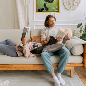 Pu&ograve; includere: Una coppia si rilassa su un divano, leggendo libri. La donna &egrave; sdraiata con la testa appoggiata sulle gambe dell'uomo. L'uomo indossa dei cerotti per gli occhi e la donna indossa una felpa rosa. Il divano &egrave; di colore marrone chiaro con gambe in legno. Lo sfondo &egrave; una parete bianca con un dipinto astratto verde e nero.