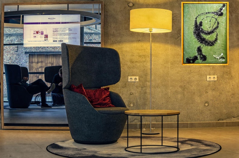 Pu&ograve; includere: Una poltrona grigia con un cuscino rosso si trova in una hall moderna con una lampada da terra, un tavolino rotondo e un dipinto astratto incorniciato sul muro.