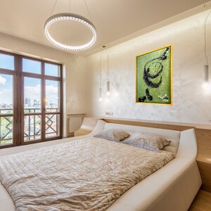 Pu&ograve; includere: Un letto bianco con un piumino beige e due cuscini in una camera da letto moderna con una grande finestra con vista sullo skyline della citt&agrave;. Un dipinto astratto verde e nero incorniciato &egrave; appeso al muro sopra il letto.