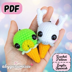 May include: Two crocheted ice cream cone-shaped amigurumi toys. One is green with a yellow beak, the other is white with bunny ears. Both have black eyes and are on yellow cones. The image includes the text "PDF", "abygurumiss", and "Crochet Pattern Englis Spanish".