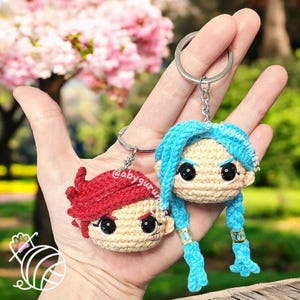 May include: Two crocheted keychain figures with blue and red hair. The figures have large eyes and are attached to silver keychains.