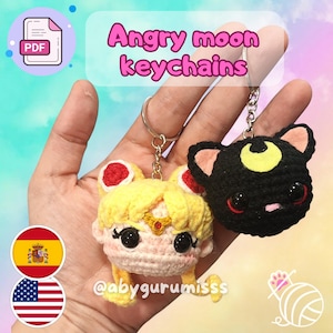 Puede incluir: Dos llaveros de ganchillo hechos a mano. Uno es un personaje amarillo con detalles rojos y una corona dorada. El otro es un gato negro con una luna creciente amarilla. El texto "Angry moon keychains" se muestra en rosa.