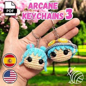 Puede incluir: Dos llaveros hechos a mano de ganchillo con caras de personajes de dibujos animados con pelo azul y ojos negros grandes. Los llaveros están sujetos a anillos de metal plateado. El texto "ARCANE KEYCHAINS 3" se muestra en la parte superior de la imagen.