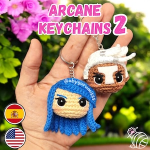 Puede incluir: Dos llaveros de ganchillo con personajes de Arcane. Un llavero es una mujer con el pelo azul y ojos marrones. El otro llavero es un hombre de piel morena con el pelo blanco y ojos marrones. Los llaveros están unidos a llaveros plateados.
