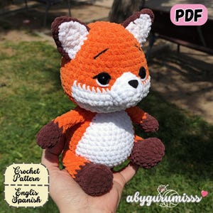 Puede incluir: Un juguete de zorro de ganchillo naranja y blanco hecho a mano con detalles marrones. El zorro tiene ojos negros y una barriga blanca. La imagen incluye el texto "PDF", "Crochet Pattern" y "English Spanish".