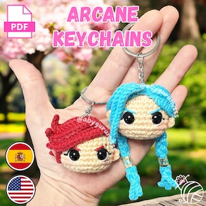 Puede incluir: Dos llaveros de personajes de crochet, uno con cabello rojo y otro con cabello azul, colgando de llaveros plateados. Los llaveros están etiquetados como "Arcane Keychains" y la imagen incluye un icono PDF y banderas de España y Estados Unidos.
