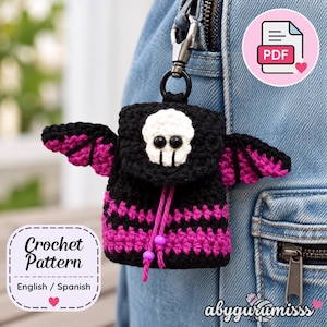 Puede incluir: Un dije de mochila tejido a crochet en negro y magenta con un motivo de calavera blanca en la solapa, ojos de botón negros y dos cordones de ajuste magenta con cuentas moradas. El artículo está sujeto a un gancho de mosquetón de metal plateado.