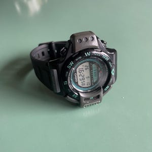 Op de afbeelding: Een zwart digitaal polshorloge met een ronde wijzerplaat en een zwarte kunststof horlogeband. Het horloge heeft een kompasring met windstreken, een digitaal display met de tijd 16:29:58, de datum 9-10 en de tekst CASIO en TRIPLE SENSOR.