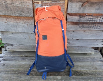 Könnte beinhalten: Ein orangefarbener und blauer Rucksack mit verstellbaren Trägern. Der Rucksack hat ein großes Hauptfach und eine kleinere Tasche vorne.