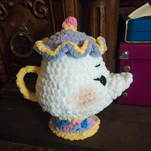 May include: A white, plush teapot with a smiling face, black eyes, and a curved spout. The teapot has a yellow handle and a decorative hat with pink, yellow, and purple accents. The base of the teapot features a colorful, patterned design.