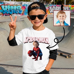 May include: A young child wearing a white Spider-Man t-shirt, black sunglasses, and a black baseball cap. The shirt features the Spider-Man logo and a picture of a child dressed as Spider-Man. The child is smiling and giving a peace sign.