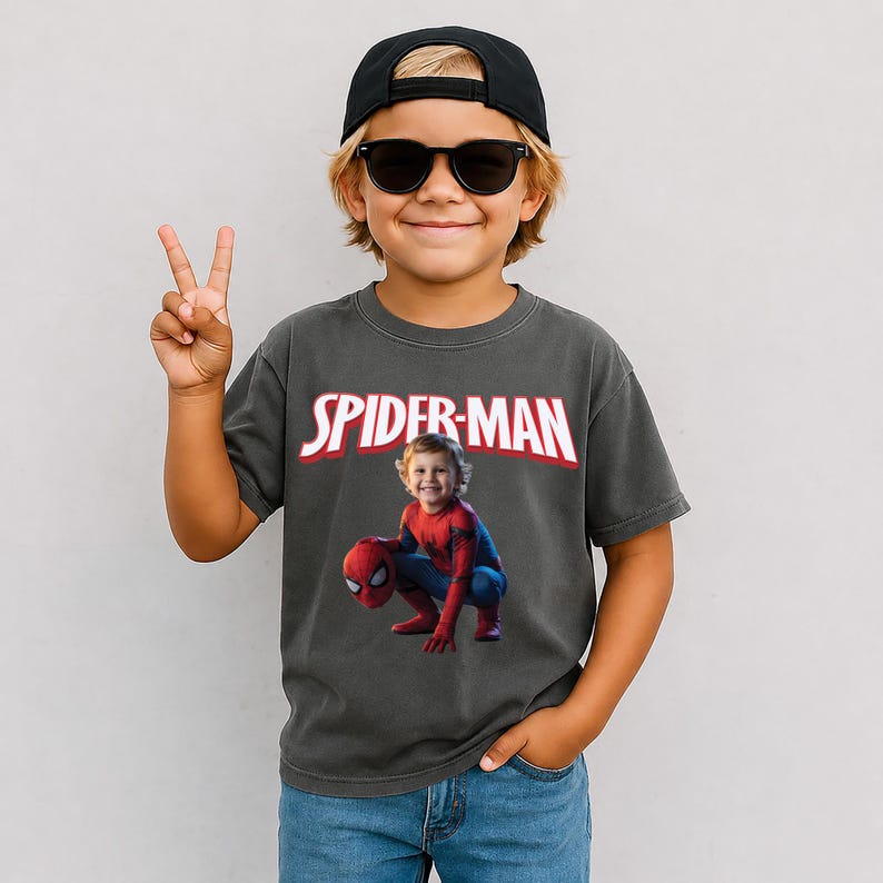 May include: A child wearing a black baseball cap and sunglasses, making a peace sign. The child is wearing a gray t-shirt with the words "SPIDER-MAN" in white and red, and a photo of a child dressed as Spider-Man.