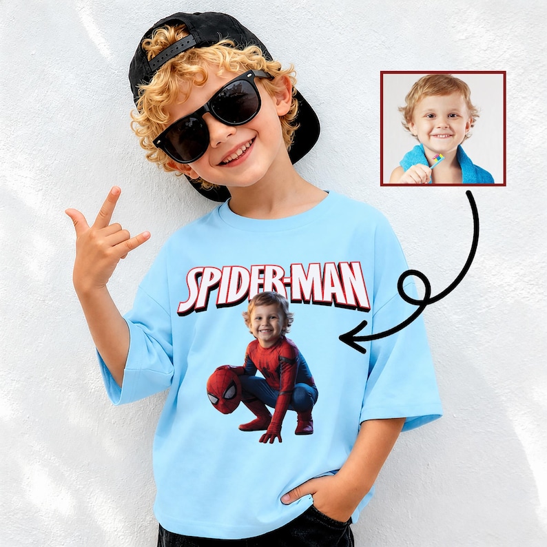 May include: A light blue Spider-Man t-shirt with the word "SPIDER-MAN" in red and white lettering. The shirt features a graphic of a child dressed as Spider-Man. The child in the image is wearing sunglasses and a black baseball cap.