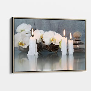 Op de afbeelding: Een ingelijst canvasprint met een stilleven van witte orchideeën, een tak drijfhout en vier brandende witte kaarsen op een grijze achtergrond. De kaarsen zijn in een rij gerangschikt en de orchideeën zijn geclusterd op het drijfhout. De afbeelding is een kalmerende en serene scène.