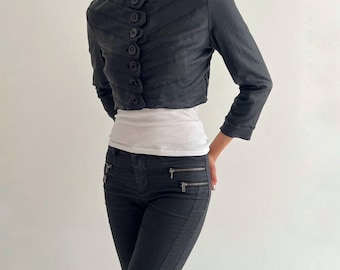 May include: A cropped black jacket featuring a high collar, three-quarter length sleeves, and a vertical row of six large, round buttons. The jacket has horizontal stitched detailing across the front and is worn over a plain white top and black pants.
