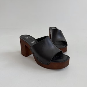 Puede incluir: Un par de mules de cuero negro con punta abierta y tacones de bloque de madera gruesos. Los zapatos cuentan con una suela de plataforma, tachuelas de metal oscuro en los laterales y la marca Bata con el texto Italian Design en la plantilla.
