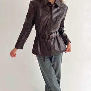 May include: A dark brown long-sleeved jacket with a pointed collar, a front button closure, and a matching belt with a circular buckle at the waist. The jacket is paired with grey denim jeans, creating a structured look with visible seams.
