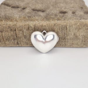 May include: A silver-colored heart-shaped pendant with a loop at the top for hanging. The pendant has a smooth, reflective surface and is set against a textured wooden background.