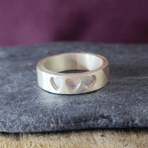 May include: A silver band ring with three heart cutouts. The ring is on a grey surface.