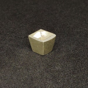 May include: A small, square, silver-colored crucible with a slightly rounded bottom.