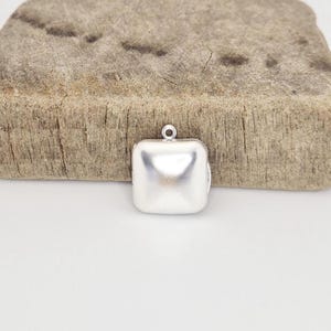 May include: A small, square, silver-colored pendant with a loop at the top. The pendant has a slightly rounded shape and a smooth, reflective surface. It is set against a light-colored background.