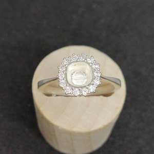 May include: A silver ring with a square-shaped setting surrounded by small clear stones. The ring is on a wooden display stand.