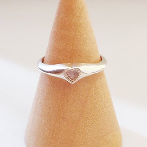 May include: A silver ring with a heart-shaped design. The ring is on a wooden ring display stand.