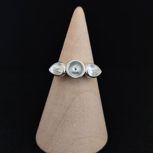 Puede incluir: Un anillo de plata con tres engastes redondos de fondo abierto. El anillo se muestra en un soporte de madera.