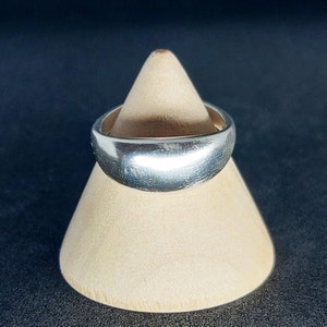 Könnte beinhalten: Ein silberner Ring mit abgerundeter, glatter Oberfläche. Der Ring wird auf einem hölzernen Ringständer präsentiert.