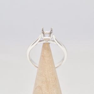 May include: A silver ring with a square setting, displayed on a wooden cone. The ring's band tapers towards the setting, creating a classic and elegant design. The setting has four prongs to hold a gemstone.