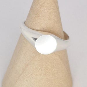 May include: A silver signet ring with a smooth, rounded band and a circular face. The ring is displayed on a light brown, cone-shaped object. The ring's design is simple and modern, suitable for everyday wear.