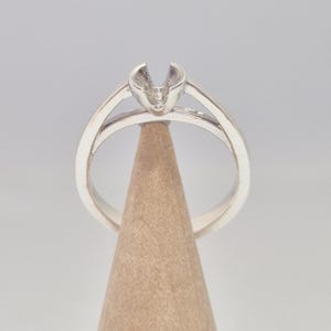 Könnte beinhalten: Ein silberner Ring mit geteiltem Schaft-Design, mit einer Krappenfassung für einen Edelstein. Der Ring wird auf einem Holzkegel vor neutralem Hintergrund präsentiert, um seine polierte Metalloberfläche hervorzuheben.