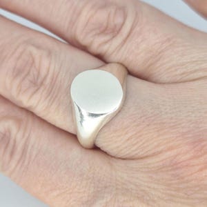 May include: A silver signet ring with a polished, circular face. The ring is shown on a hand, highlighting its simple, elegant design. The ring's band is smooth and tapers towards the back.