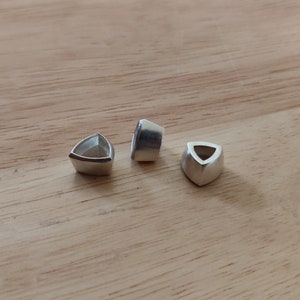 May include: Three silver triangular-shaped bezels with a hole in the center. The bezels are on a wooden surface.