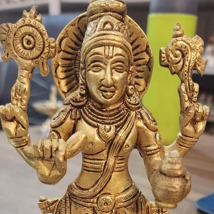 Może przedstawiać: Złota metalowa statuetka hinduskiego bóstwa z czterema ramionami, trzymająca kwiat lotosu, muszlę, dysk i dzban. Statuetka stoi na kwadratowej podstawie.