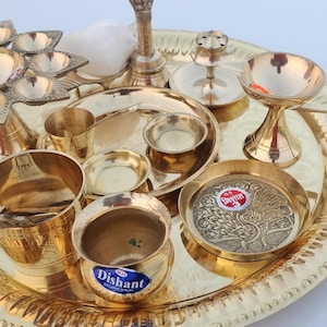Op de afbeelding: Een messing puja thali-set met verschillende items, waaronder een diya, een kalash, een lota en een klein bord. De thali is versierd met een verhoogde rand en een bloemmotief.