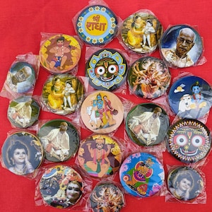 Op de afbeelding: Een verzameling ronde, kleurrijke knopen met afbeeldingen van hindoeïstische goden en religieuze symbolen. De knopen zijn in de vorm van een hart op een rode achtergrond gerangschikt. Sommige van de afgebeelde goden zijn Krishna, Radha en Shiva.