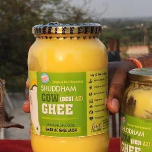 Puede incluir: Dos tarros de vidrio de ghee, una mantequilla clarificada tradicional india. El tarro más grande está etiquetado como "Shuddham Cow (Desi A2) Ghee" y el tarro más pequeño está etiquetado como "Shuddham Cow (Desi A2) Ghee". Ambos tarros tienen una etiqueta amarilla con acentos verdes.