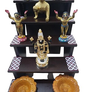 May include: A three-tiered wooden shelf with decorative details, featuring five gold and silver figurines of Hindu deities, and two ornate brown clay diyas.