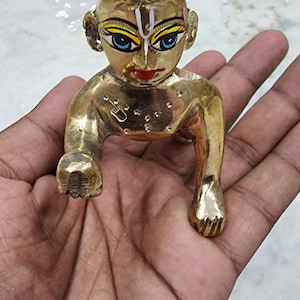 May include: A small, gold-colored statue of a baby Krishna, sitting with his legs crossed and hands raised. The statue has painted details, including blue eyes, a red mouth, and a white line on the forehead.