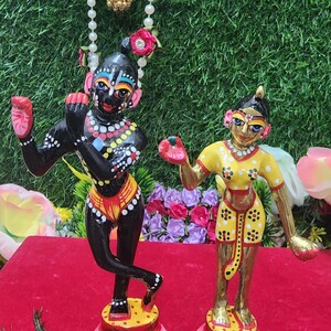Op de afbeelding: Twee handgeschilderde beeldjes van hindoe&iuml;stische goden, Radha en Krishna, die op lotusvormige bases staan. Krishna is zwart met kleurrijke details, en Radha draagt een gele jurk met witte en zwarte stippen. De beeldjes staan op een rood fluwelen oppervlak met een pauwenveer.