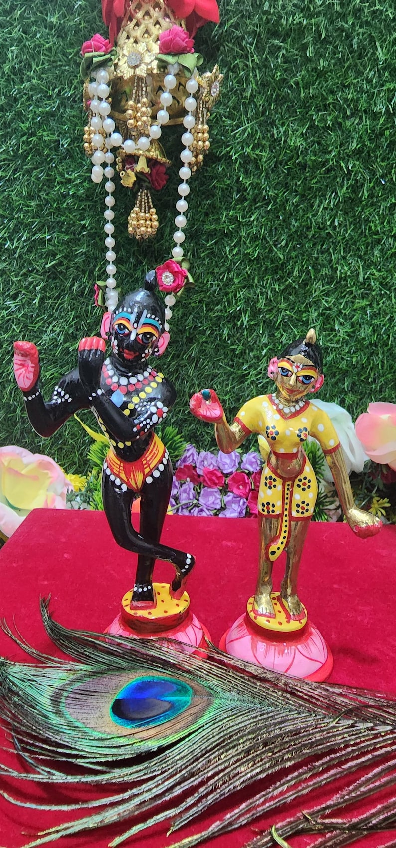 Op de afbeelding: Twee handgeschilderde beeldjes van hindoe&iuml;stische goden, Radha en Krishna, die op lotusvormige bases staan. Krishna is zwart met kleurrijke details, en Radha draagt een gele jurk met witte en zwarte stippen. De beeldjes staan op een rood fluwelen oppervlak met een pauwenveer.