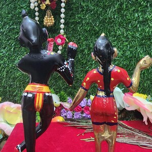 Op de afbeelding: Een paar handgeschilderde beeldjes die een stel in traditionele Indiase kleding voorstellen. Het mannelijke beeldje is zwart met gele en rode accenten, terwijl het vrouwelijke beeldje een rode jurk draagt met gele en blauwe accenten. Beide beeldjes staan op lotusvormige voetstukken.